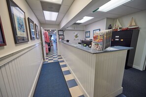 A hotel reception area with a check-in counter, framed pictures on the walls, a blue and beige checkered floor, and a cabinet with model ships.