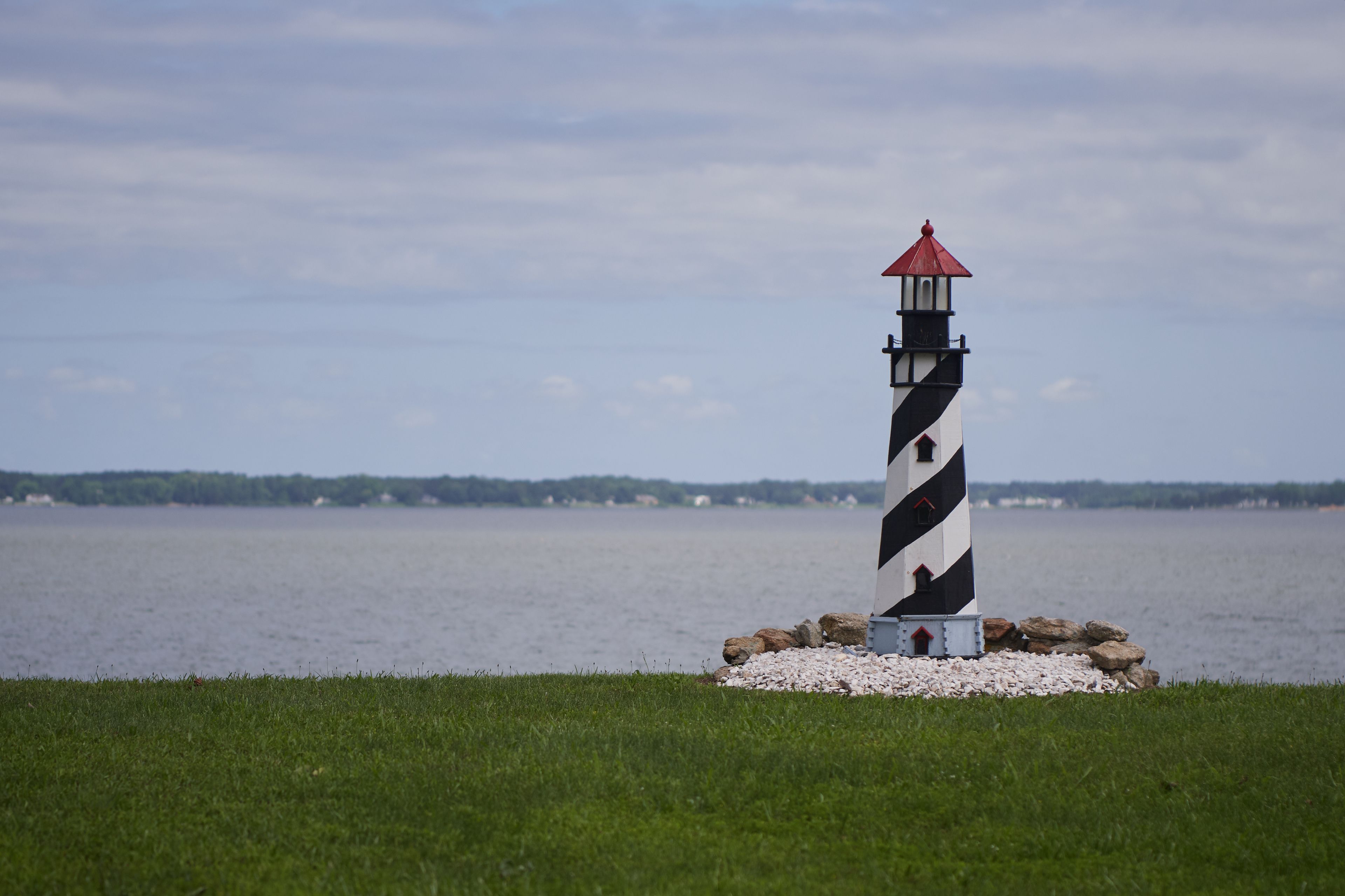 Un faro con la parte superior roja, ubicado en una zona cubierta de hierba con una masa de agua al fondo.
