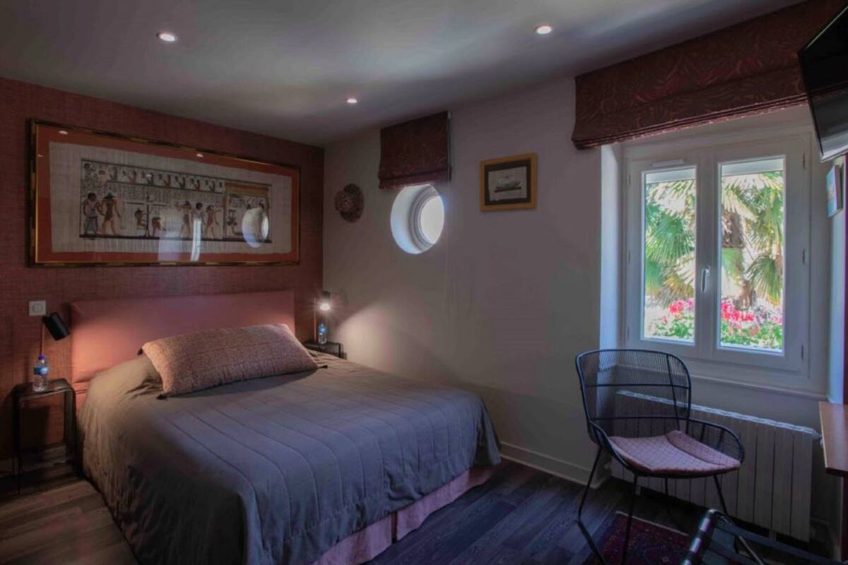 Een slaapkamer met een bed, een stoel, een raam met uitzicht op groen en een aan de muur hangend schilderij.