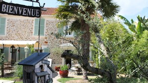A rustic wooden birdhouse with a 'WELCOME' sign, a stone building with a tiled roof, and a palm tree in the foreground. - Le Petit Nailly (Magny-les-Hameaux)