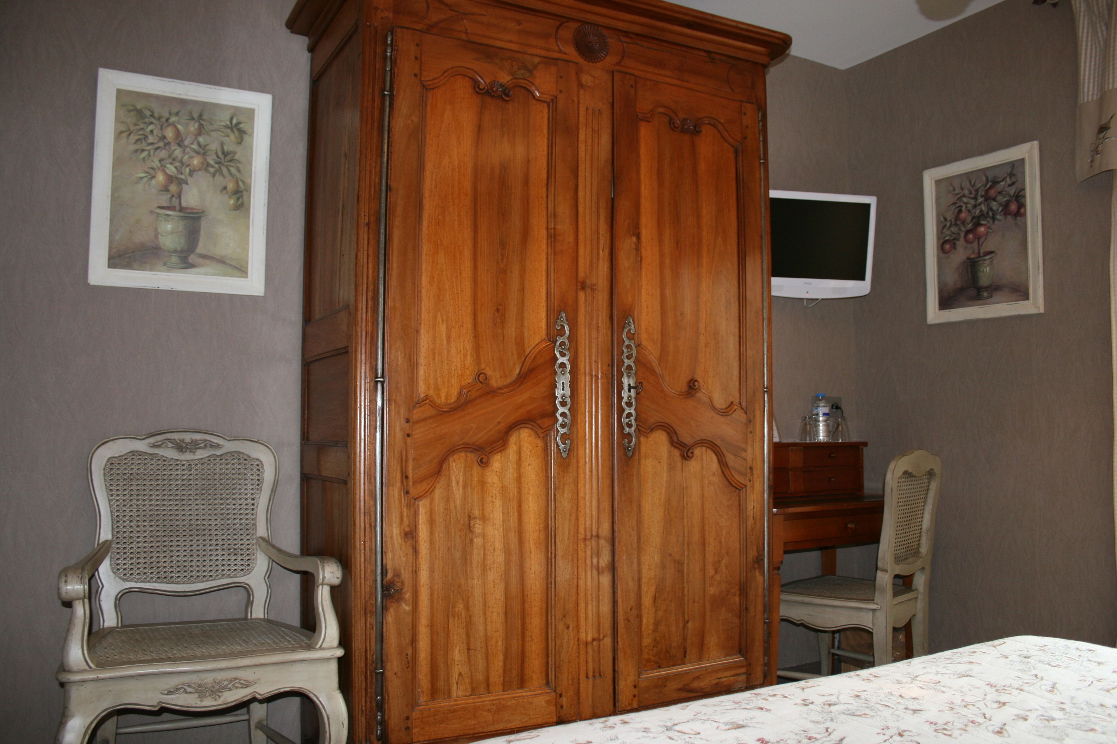 Ein Zimmer mit Holzschrank, zwei Stühlen, einem Bett mit Blumenmuster und zwei an der Wand befestigten Fernsehgeräten.