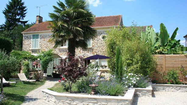 A stone house with a tiled roof, a palm tree, and a garden with various plants and a pathway. - Le Petit Nailly (Magny-les-Hameaux)