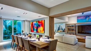 A modern dining area with a wooden table and chairs, a large abstract painting on the wall, and a living area with a sofa and TV. - Downtown Villa (Playa del Carmen)