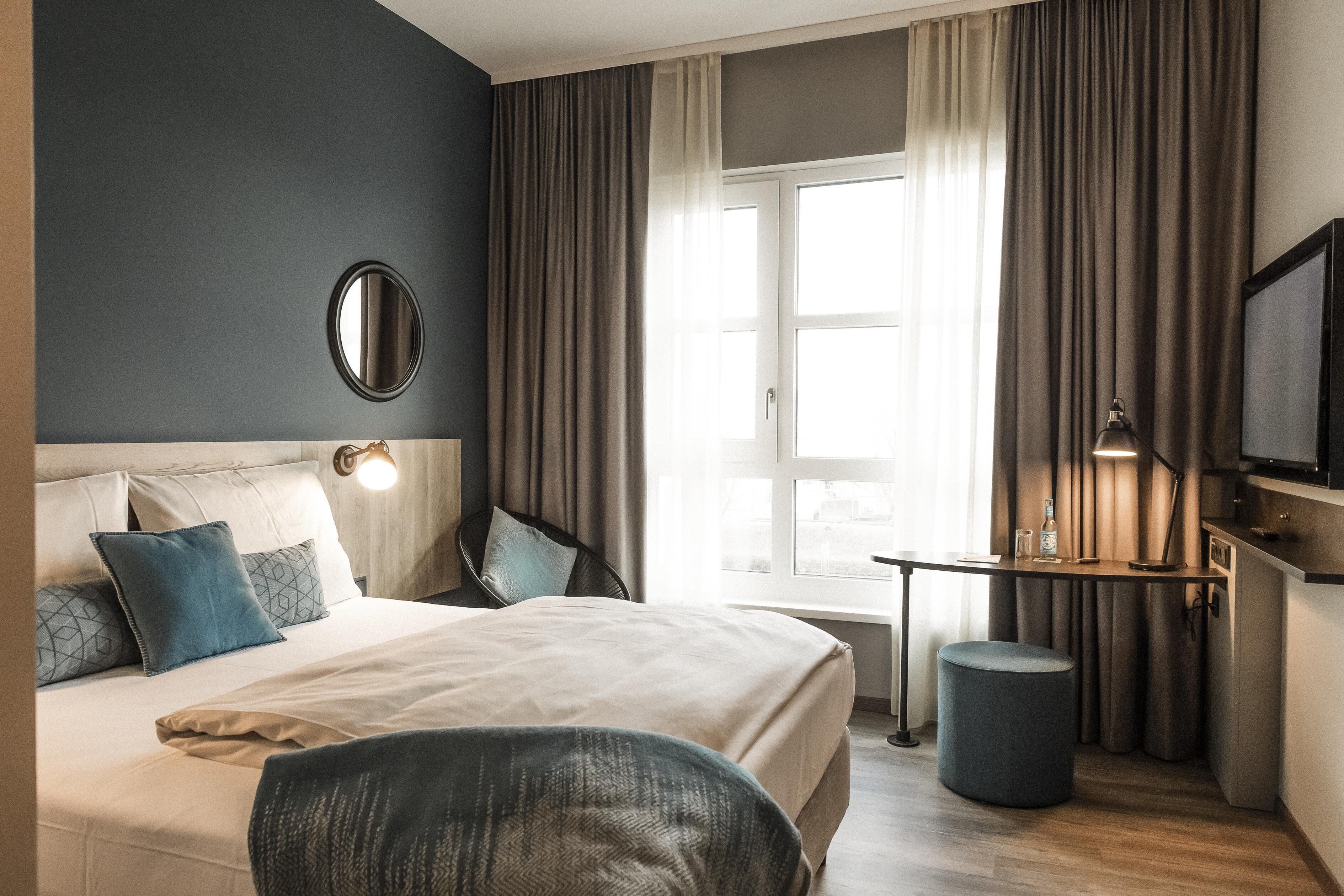 Habitación de hotel con cama, una silla, una mesita, televisión y ventana con cortinas.