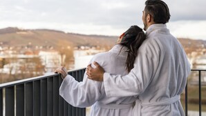 Ein Paar umarmt sich auf einem Balkon mit Blick auf eine Stadtlandschaft.