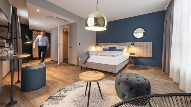 A modern hotel room with a bed, bedside table, lamp, and a round mirror on the wall. - HARBR. Hotel Heilbronn, a member of Radisson Individuals (Heilbronn)