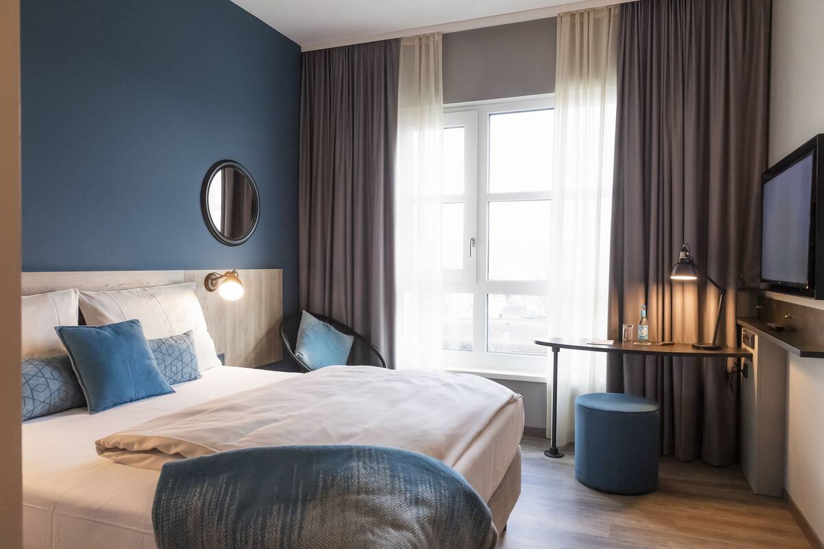 A hotel room with a bed, a desk, a chair, a round mirror, a television, and a window with curtains.