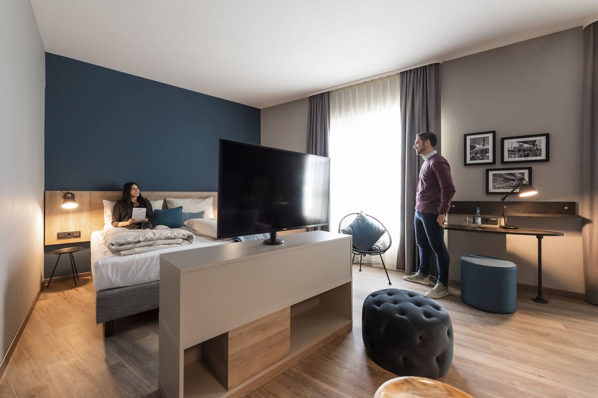 A modern hotel room with a bed, a TV, a desk, and a chair.