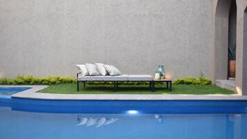 A poolside area with a lounge chair and cushions, a small table with a bottle, and a potted plant.