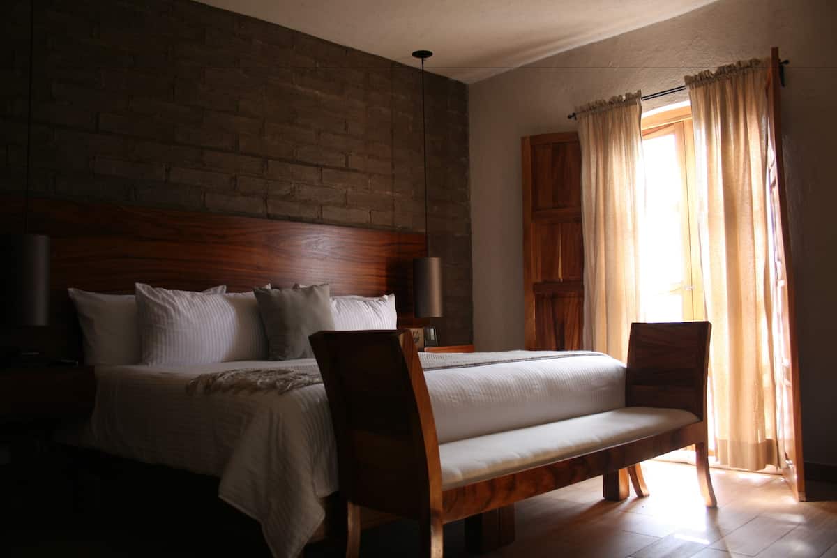 A bedroom with a brick wall, wooden headboard, a bed with white bedding, two chairs, and a window with curtains.