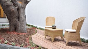Una terraza de madera con dos sillas de mimbre y una mesa pequeña, rodeada por un jardín con acolchado y plantas.