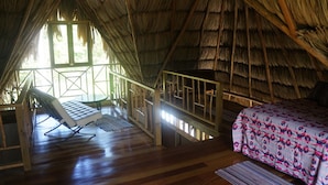 Una habitación con techo de paja, suelo de madera, cama con colcha floral, una silla y una mesita.