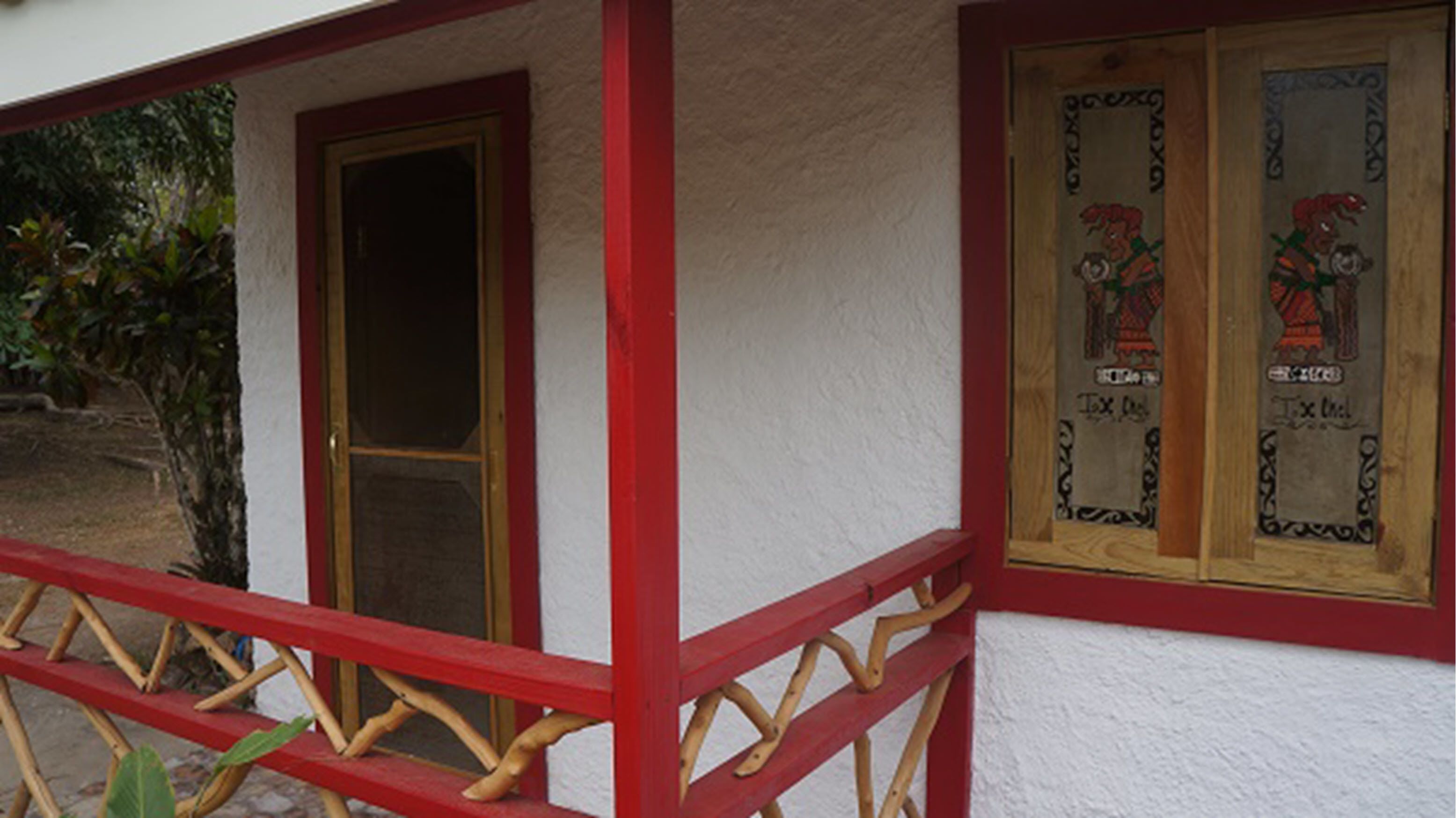 A building with a red-framed door and windows, a wooden bench, and a small tree in front.