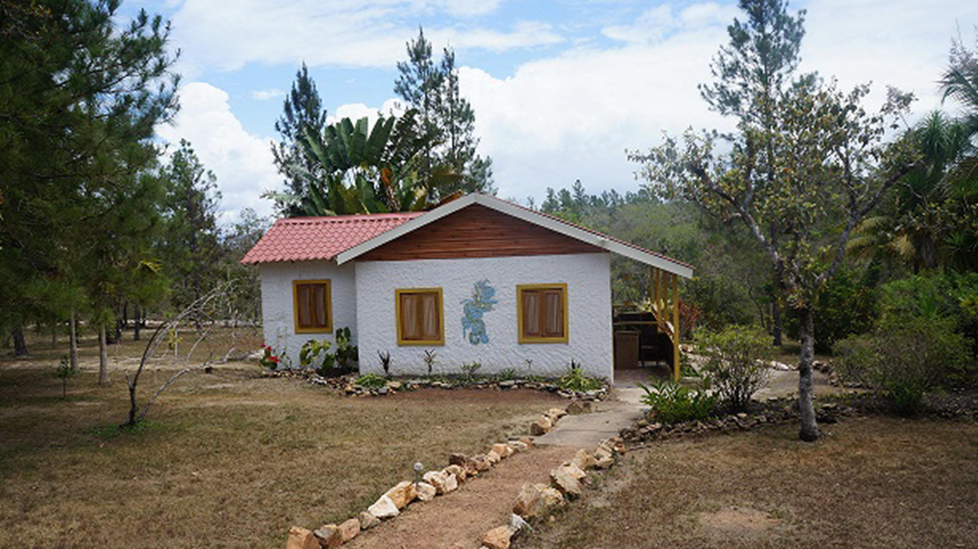 Una pequeña casa de techo rojo, paredes blancas y contraventanas de madera. Está rodeada de árboles y tiene un camino de piedra que conduce a ella.