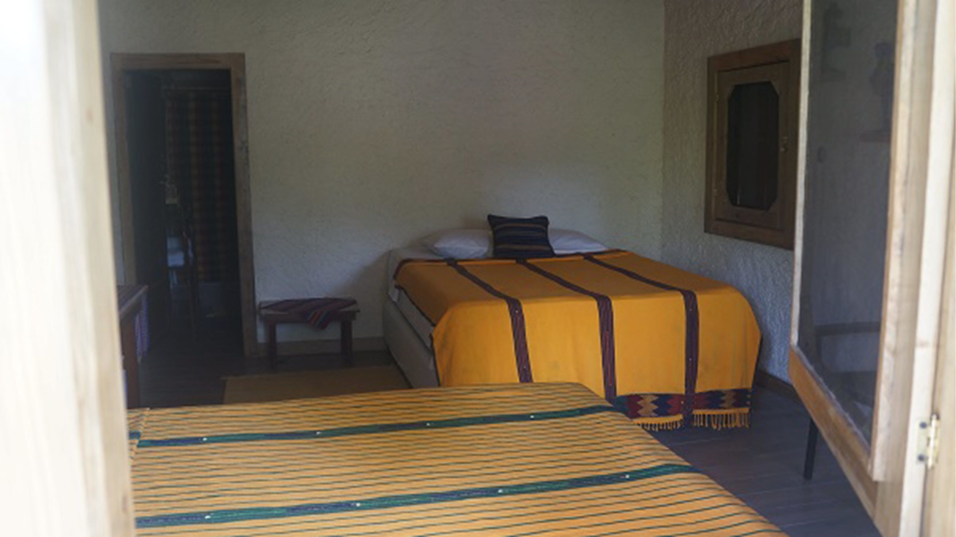 Ein Zimmer mit zwei Betten, eines mit einer gestreiften Bettdecke und das andere mit einer einfarbigen gelben Bettdecke. In der Ecke steht ein kleiner Tisch und ein Stuhl, und auf der linken Seite befindet sich eine Tür.