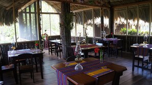 Un restaurante con mesas y sillas de madera, ventanas amplias y techo de paja.