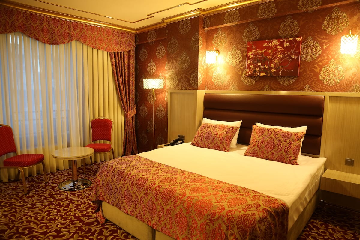 A hotel room with a large bed, two red chairs, a small round table, and a wall-mounted lamp.