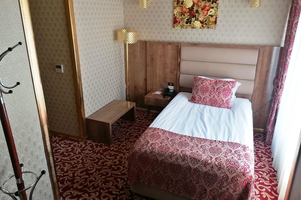A hotel room with a wooden headboard, a bed with a patterned bedspread, a bedside table with a phone and a lamp, and a floral artwork on the wall.