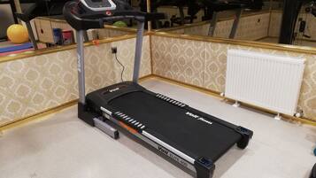Una cinta de correr Voit Elite 400 en una habitación con papel tapiz estampado, un radiador y equipo de ejercicio al fondo.