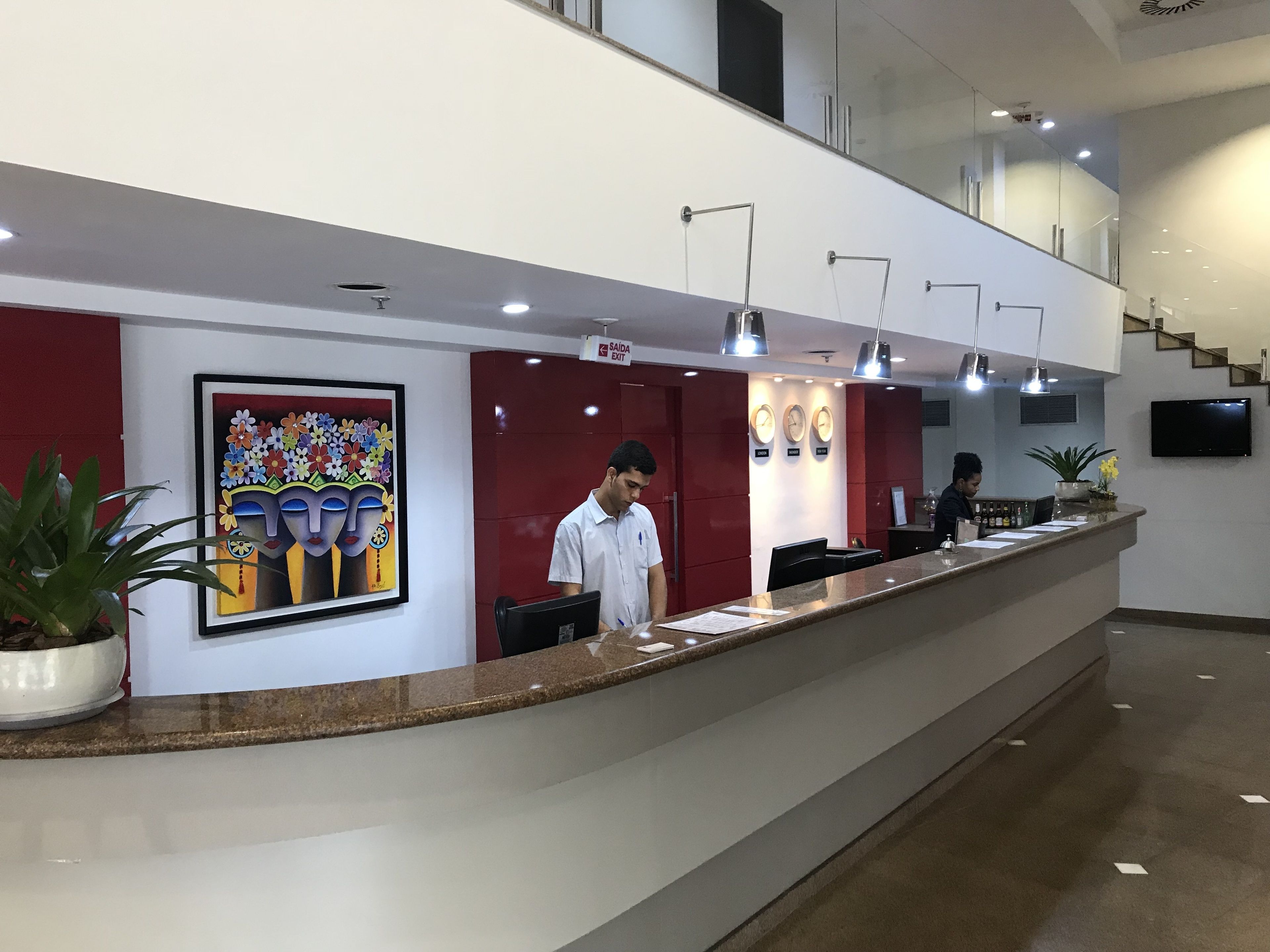Zona de recepción de un hotel con mostrador, un cuadro, relojes y una escalera.