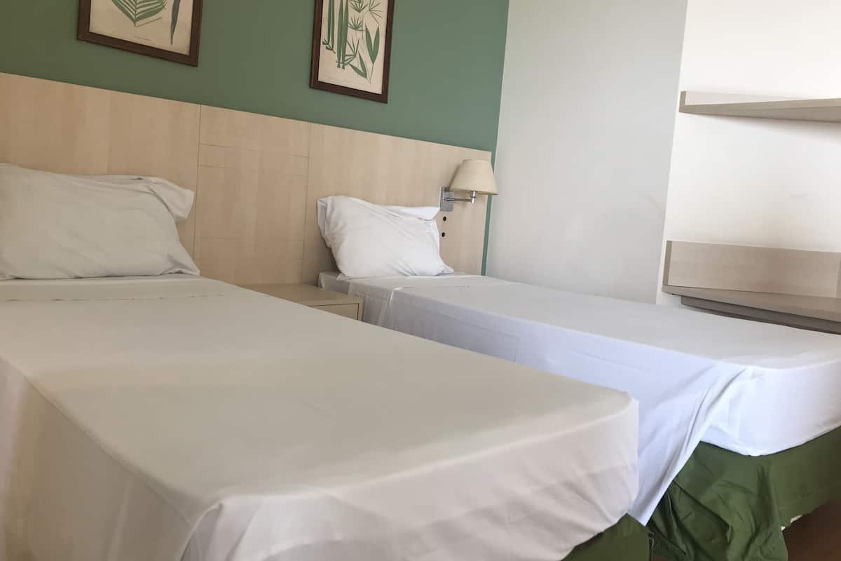 Dos camas individuales con ropa de cama blanca, un cabecero de madera, una mesita de noche con una lámpara e impresiones botánicas enmarcadas en la pared.