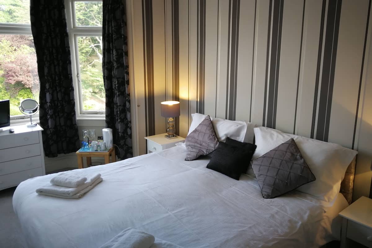 A neatly made bed with pillows, a bedside lamp, a small table with a mirror, and a window with curtains.