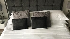 A neatly made bed with a tufted headboard, two black and one grey pillow, and two folded towels on the bed. - The Grove (Whitby)