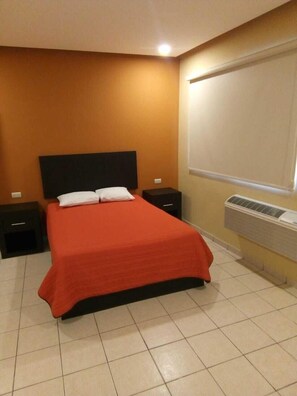 A bedroom with a bed, two nightstands, a window with blinds, and an air conditioning unit.