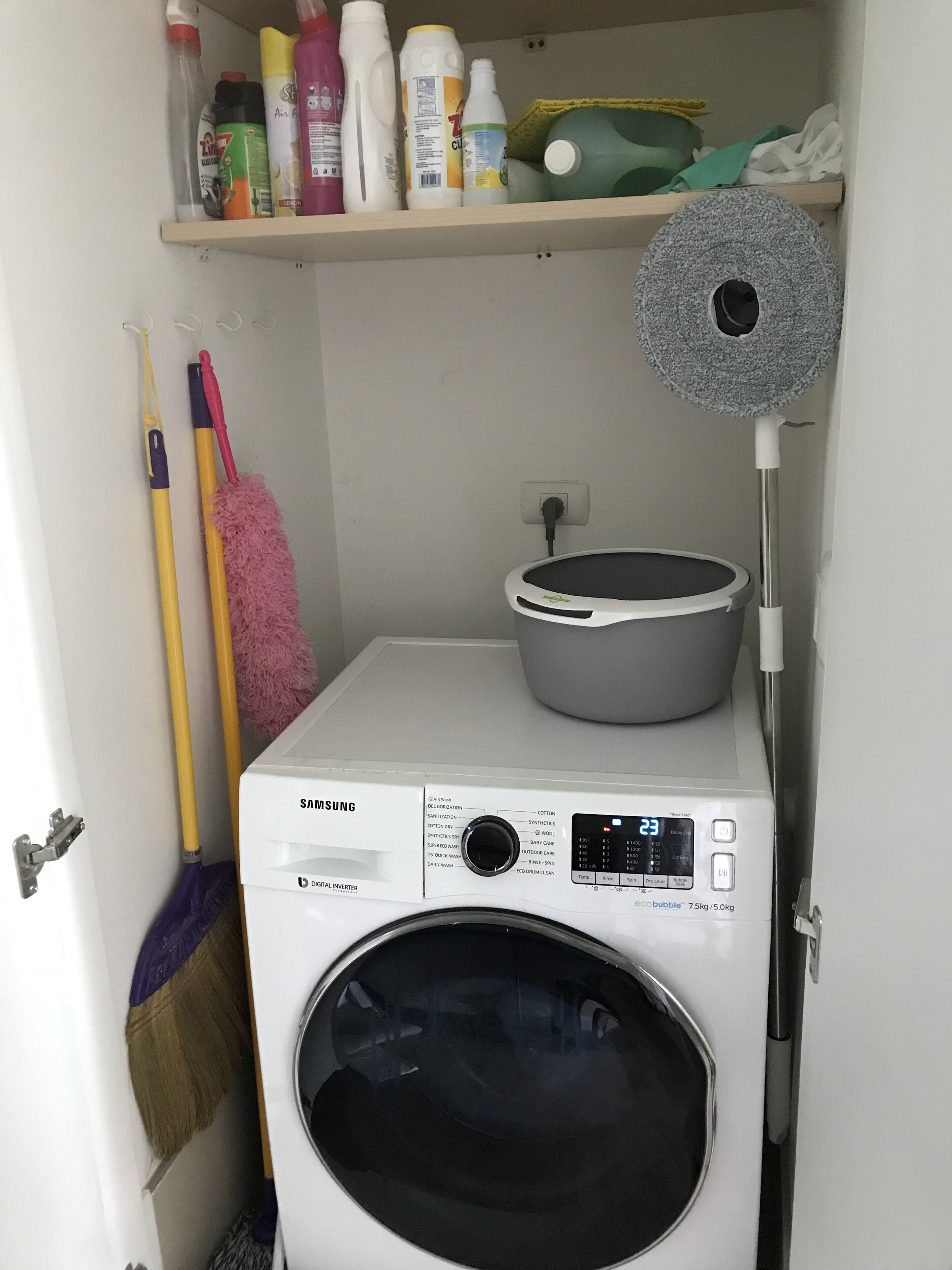 A front-loading Samsung washing machine with a digital display, a mop, and cleaning supplies on a shelf above it.