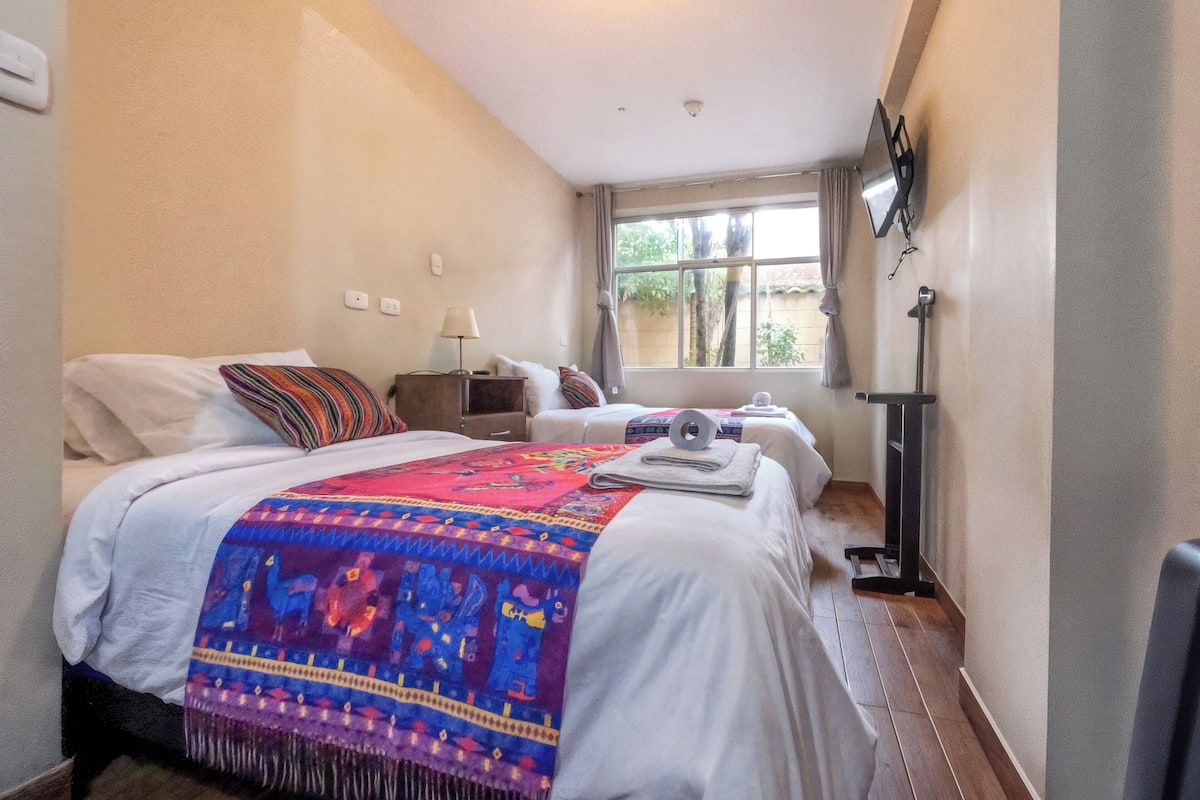 Quarto de hotel com duas camas, janela com vista para árvores, televisão de tela plana na parede e mesa de cabeceira com abajur.