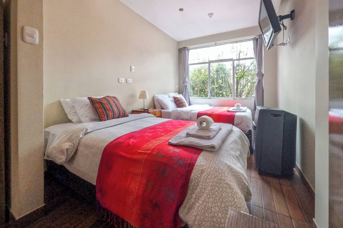 Quarto de hotel com duas camas, uma televisão, uma janela com vista para árvores e uma luminária no teto.