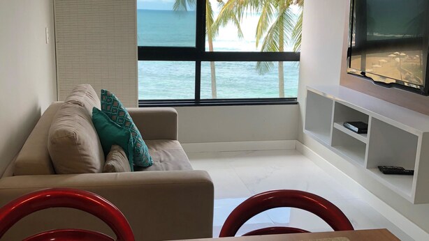 A modern living room with a view of the ocean, a sofa with cushions, a TV mounted on the wall, and a kitchen area with a gas stove. - Ondina Apart Hotel (Salvador)