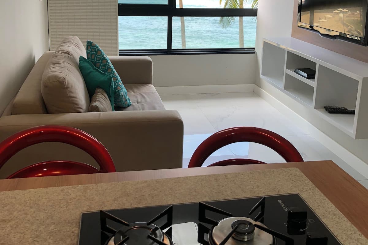 Un salon moderne avec vue sur l’océan, un canapé avec des coussins, un téléviseur fixé au mur et un coin cuisine équipé d’une cuisinière à gaz.