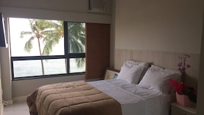 A bedroom with a bed, a window with a view of palm trees and the sea, a bedside table with a vase of flowers, and a wall-mounted air conditioner. - Ondina Apart Hotel (Salvador)