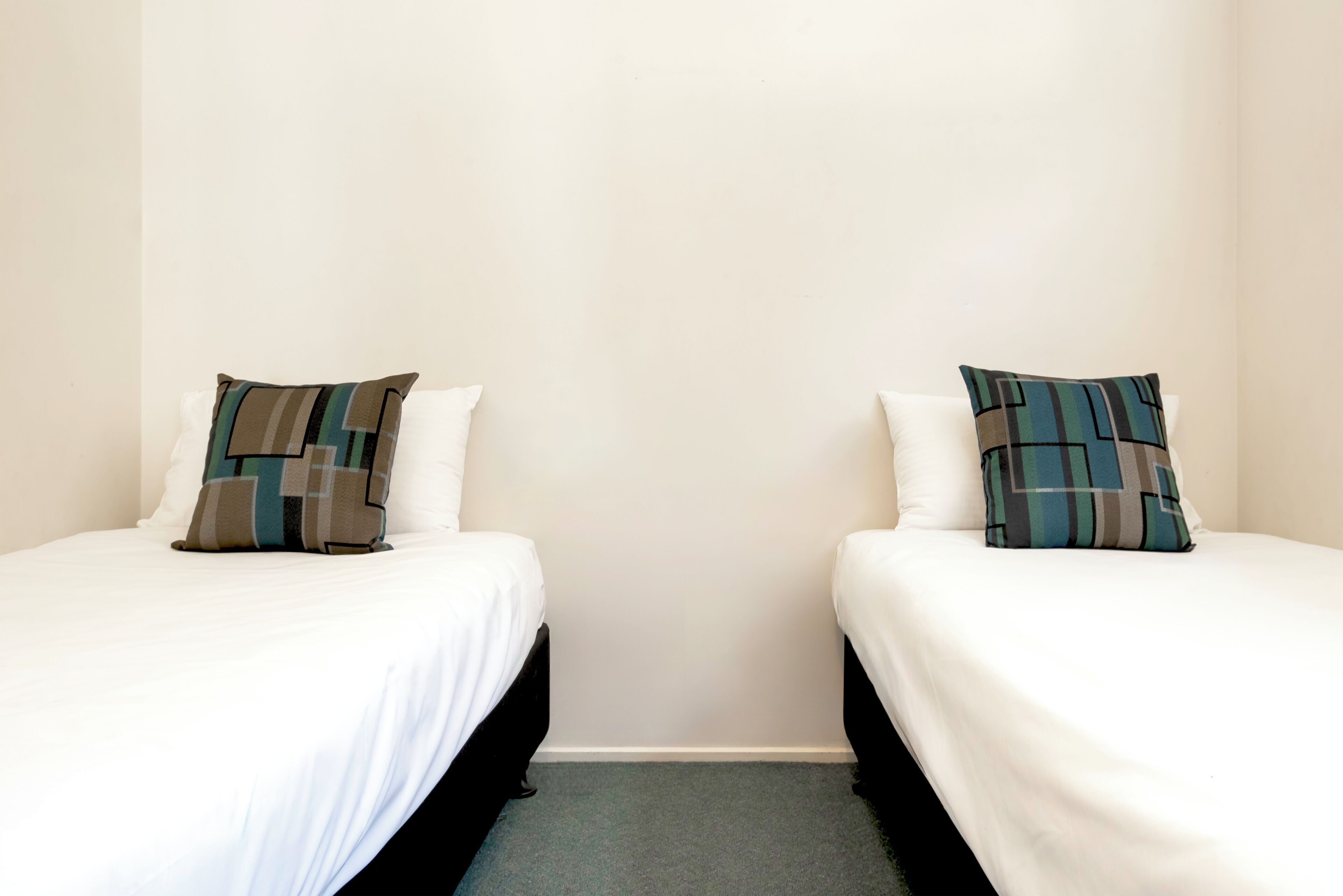 Two single beds with white linens and patterned pillows, a plain wall, and a carpeted floor.
