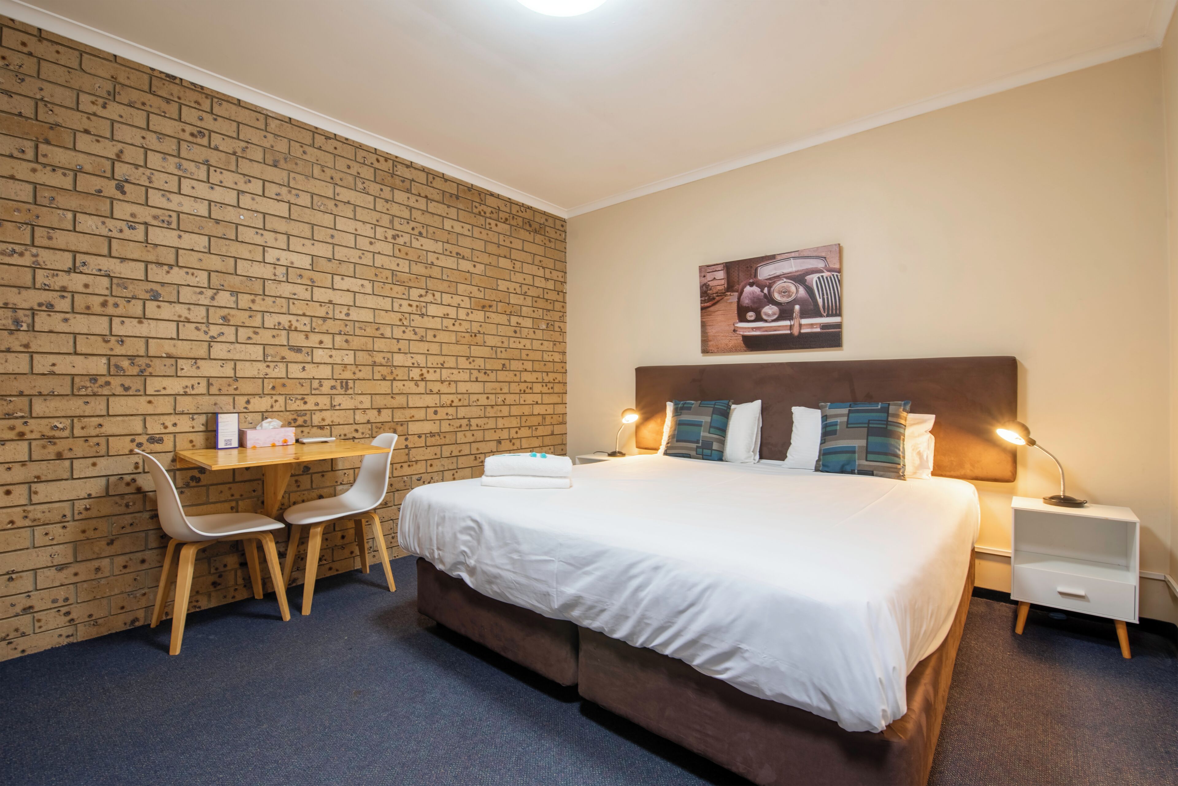 Une chambre d’hôtel avec un mur en briques, un lit avec une literie blanche, deux tables de chevet avec des lampes, un bureau avec deux chaises et un tableau accroché au mur.