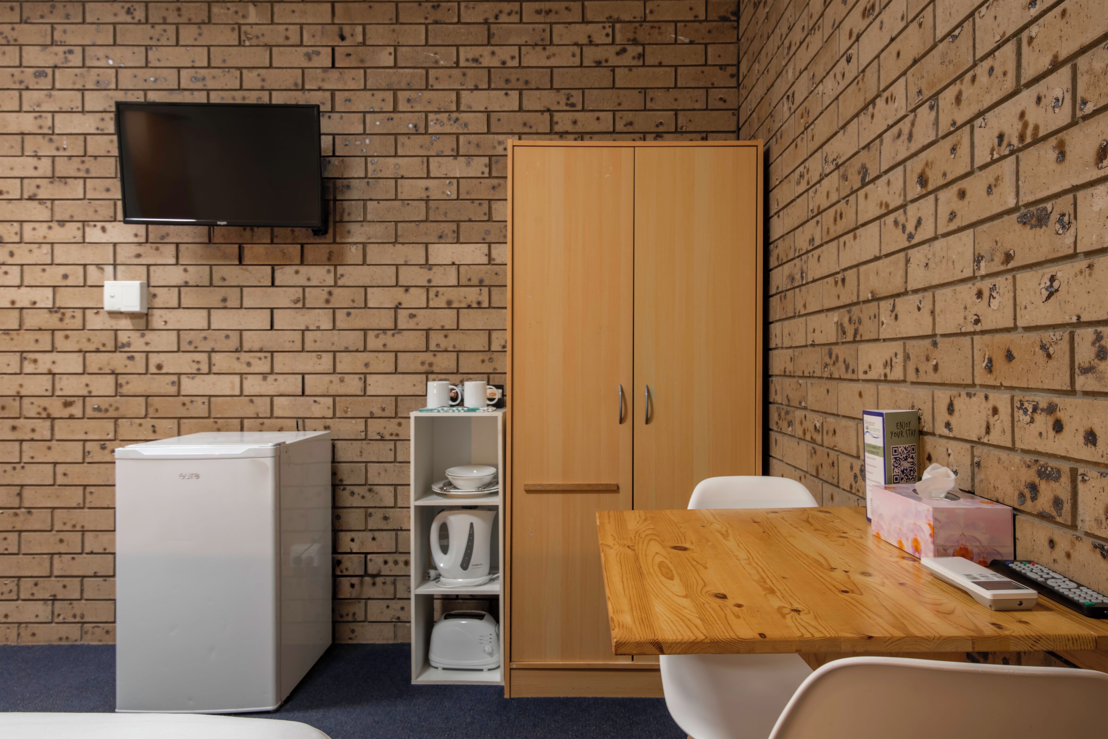 A small room with a brick wall, a TV mounted above a white refrigerator, a wooden cabinet, and a table with a remote control.