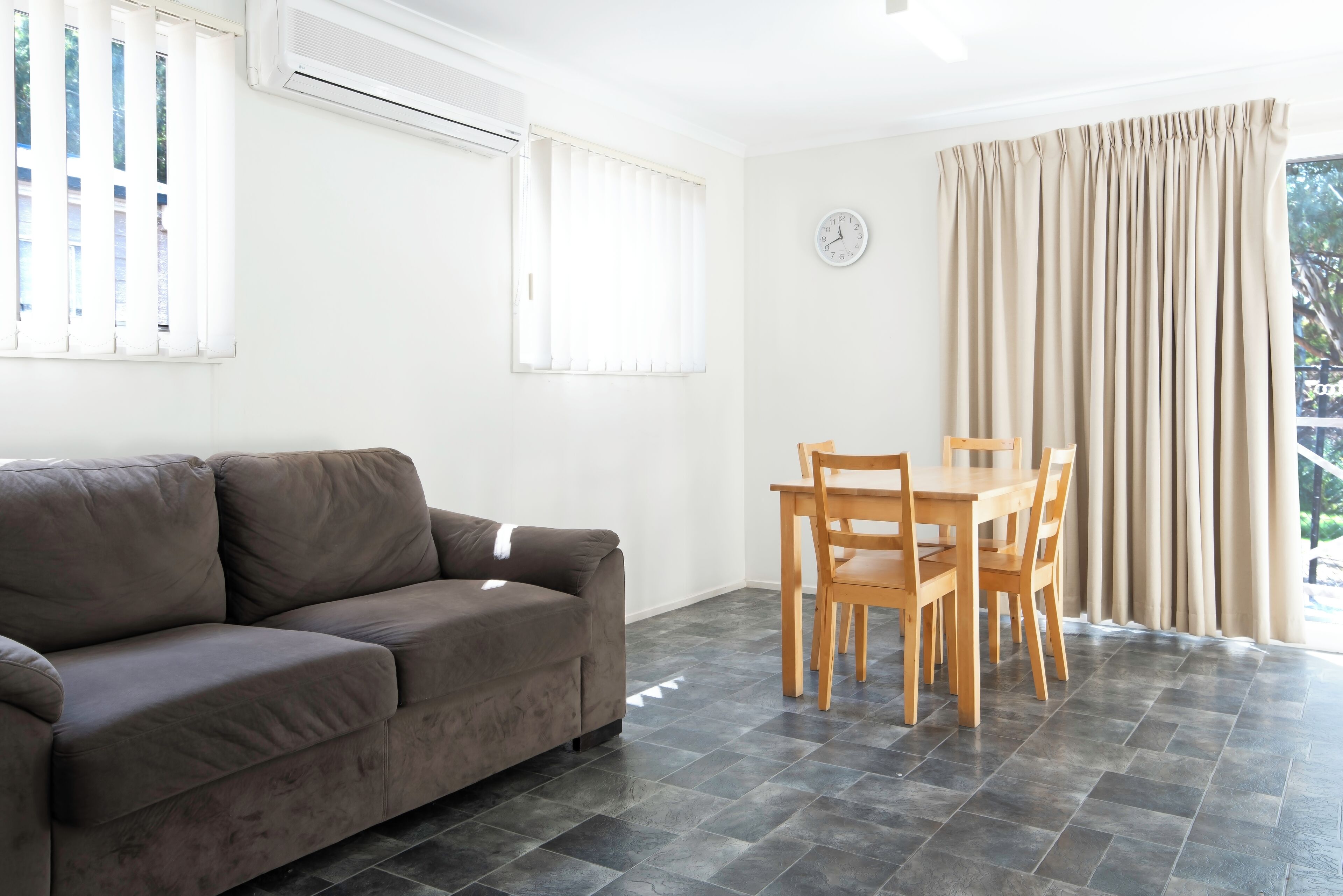 Een woonkamer met een bruine bank, een houten eettafel met stoelen, een klok aan de muur en een raam met jaloezieën.