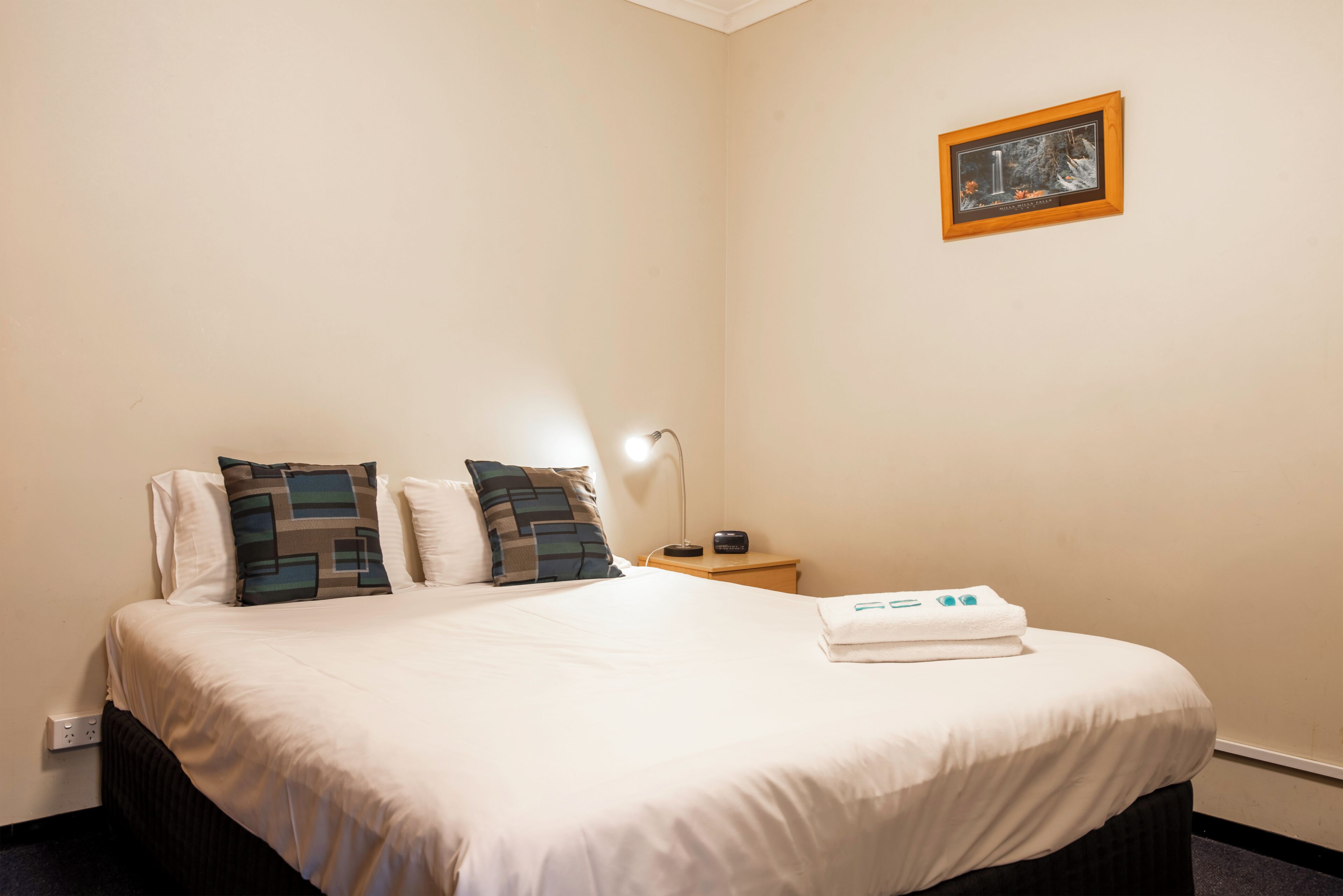 A neatly made bed with white linens and two decorative pillows, a bedside table with a lamp, and a framed picture on the wall.