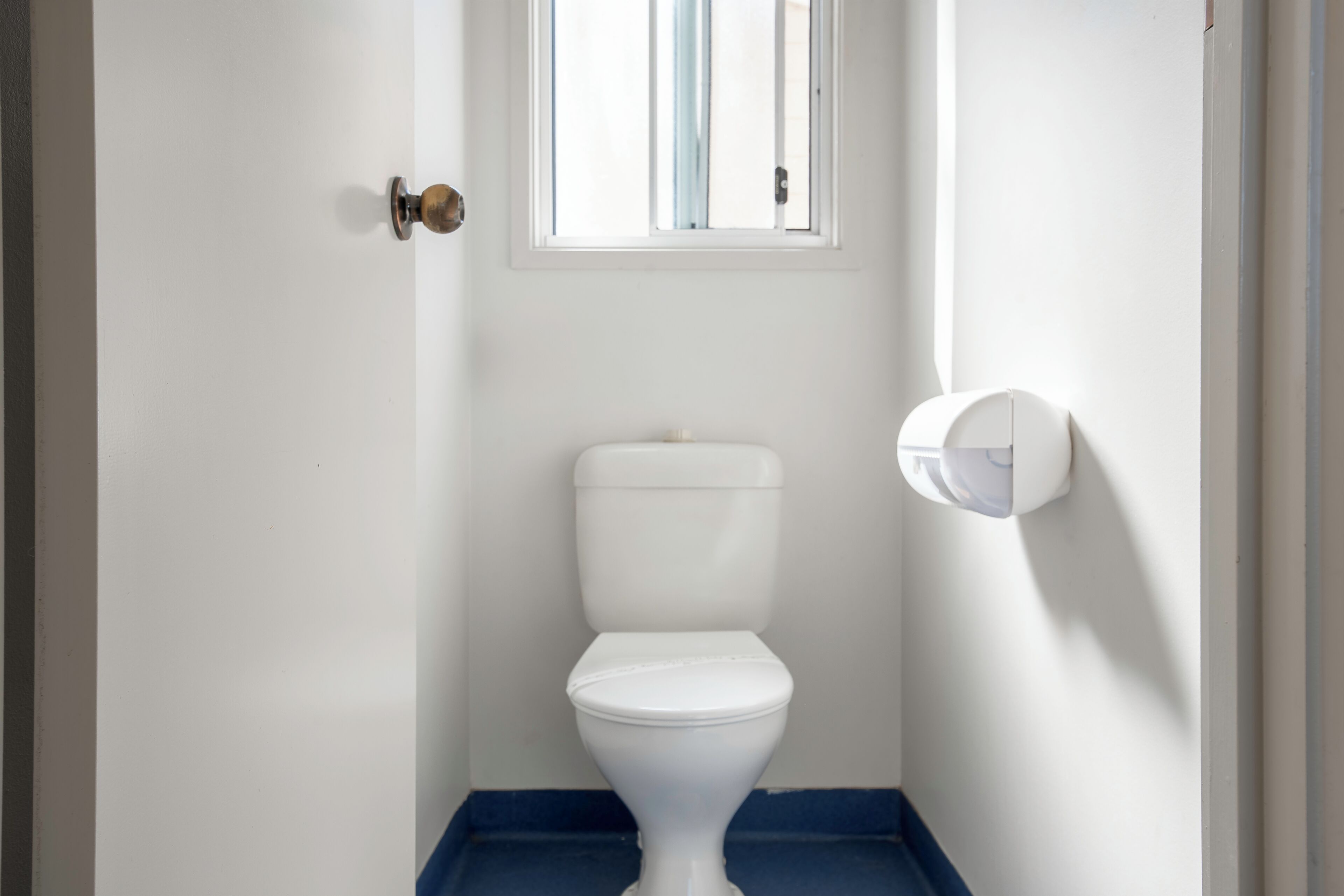 Un inodoro pequeño y blanco con base azul en un cuarto estrecho con una ventana y una puerta.