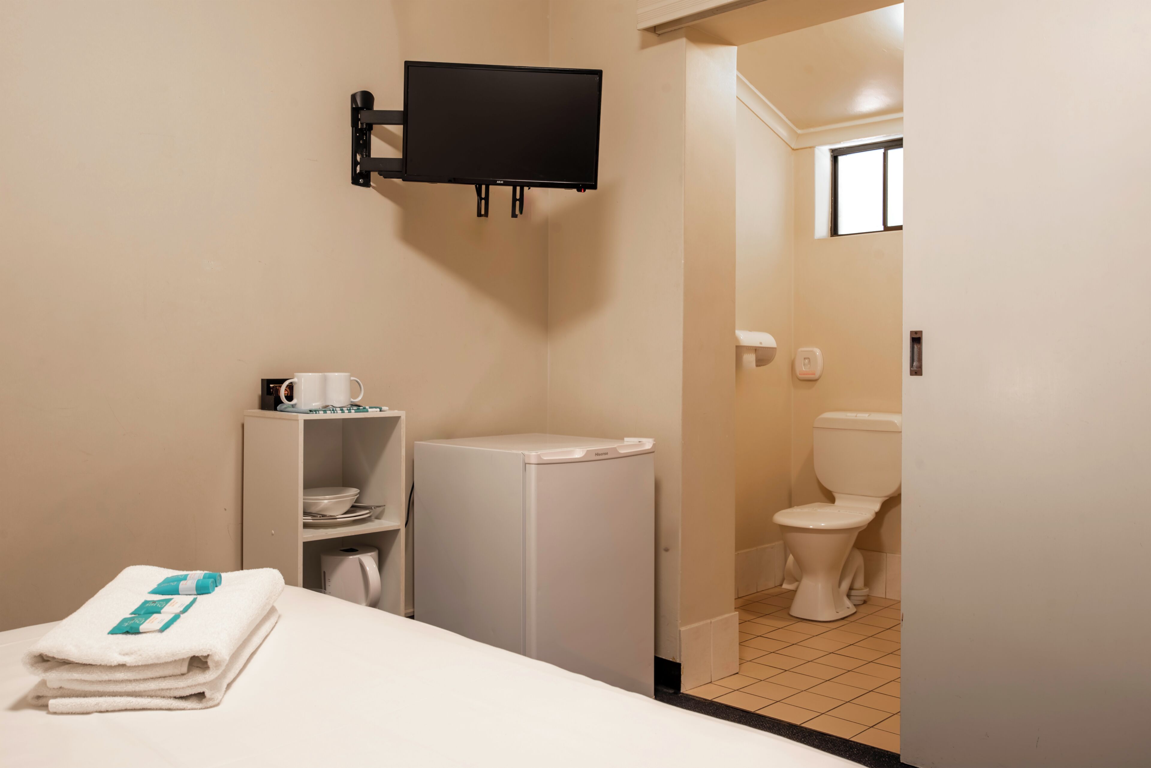 A hotel room with a bed, a small white refrigerator, a mounted TV, a white toilet, and a bathroom with a window.