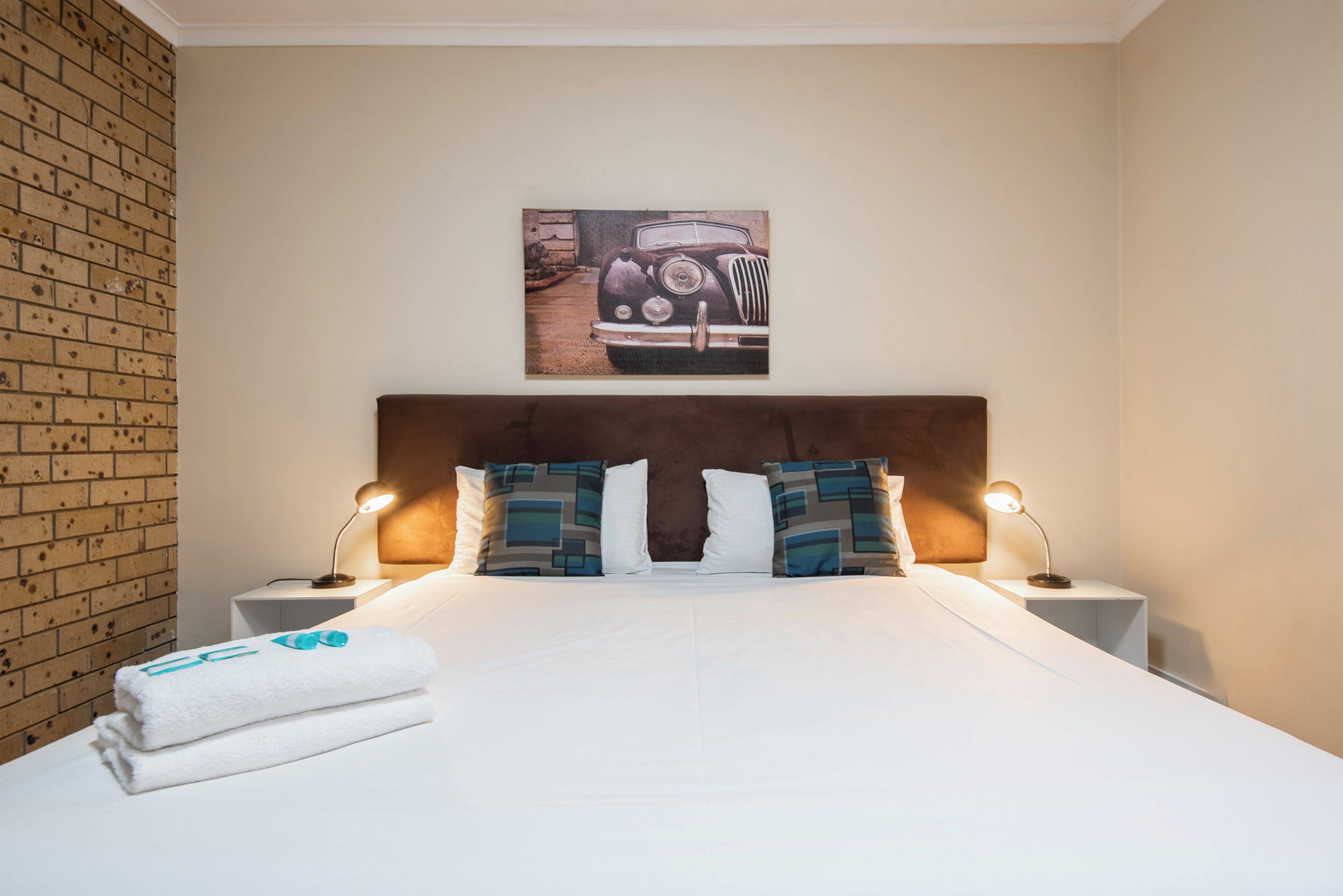 Een hotelkamer met een bakstenen muur, een bed met witte lakens, twee nachtlampjes en een schilderij van een oldtimer.