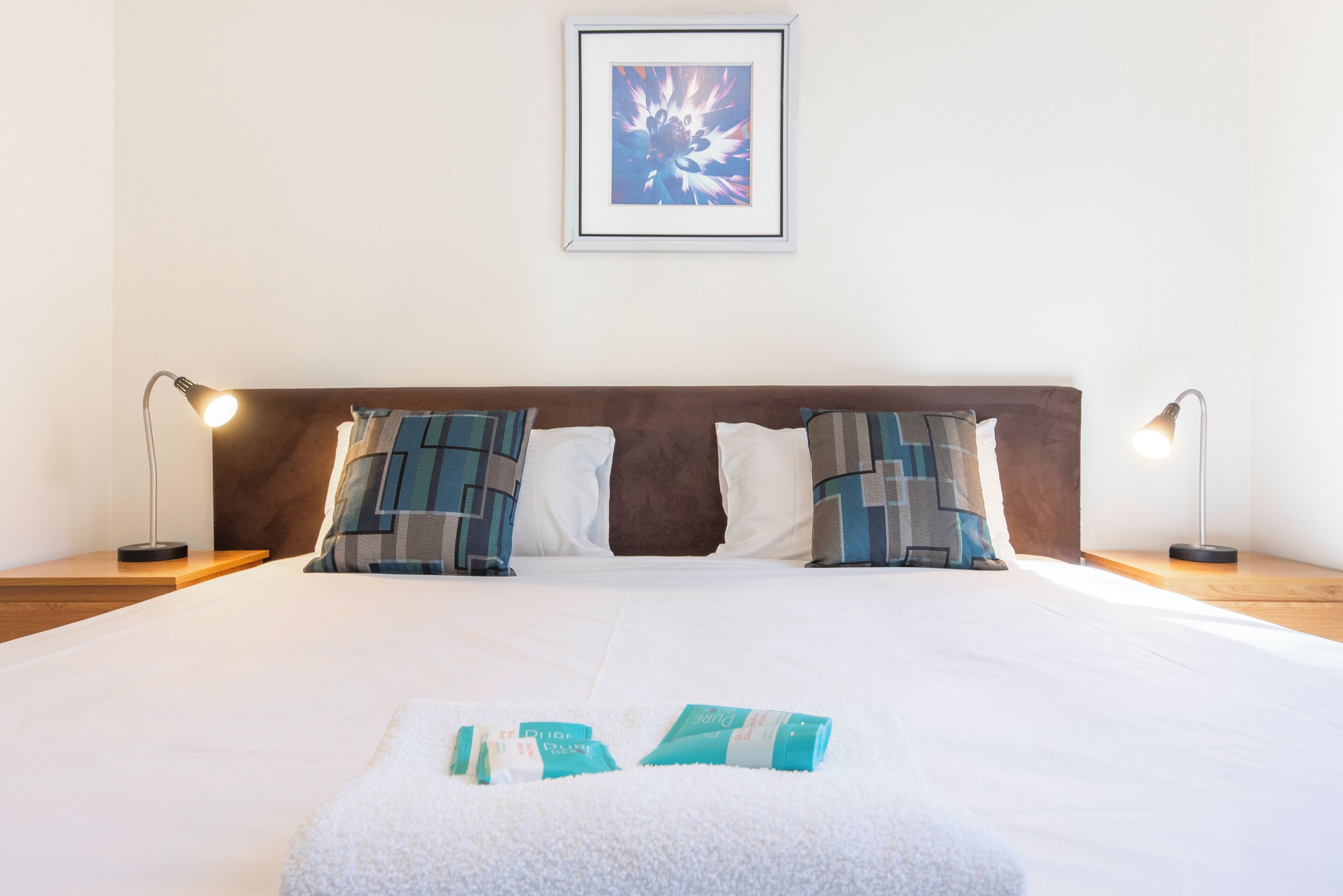 A neatly made bed with two pillows, a framed picture on the wall, and two bedside lamps.