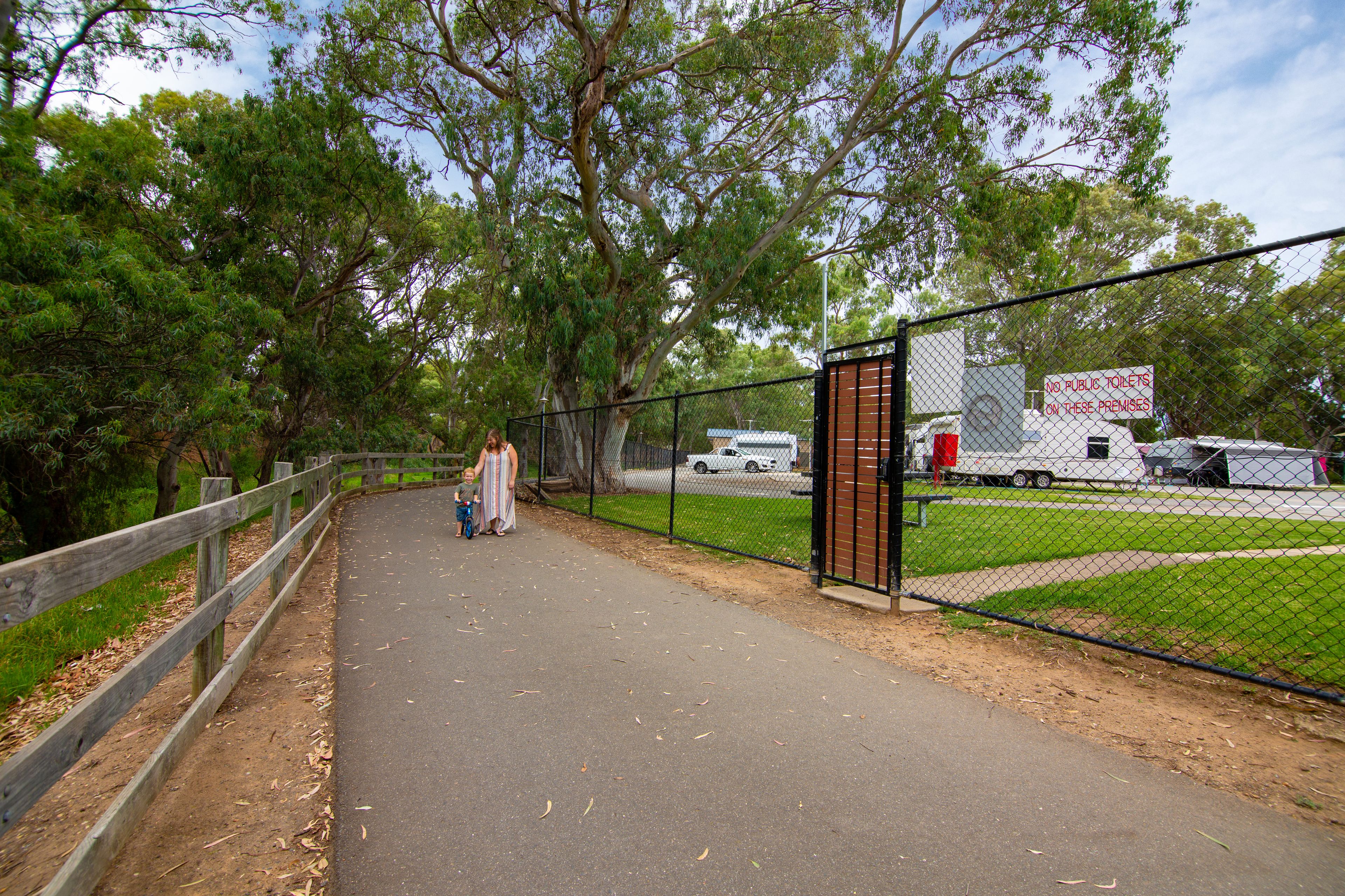 A paved walkway with a wooden fence on one side and a chain-link fence on the other, leading to a gated area with a sign prohibiting public toilets.
