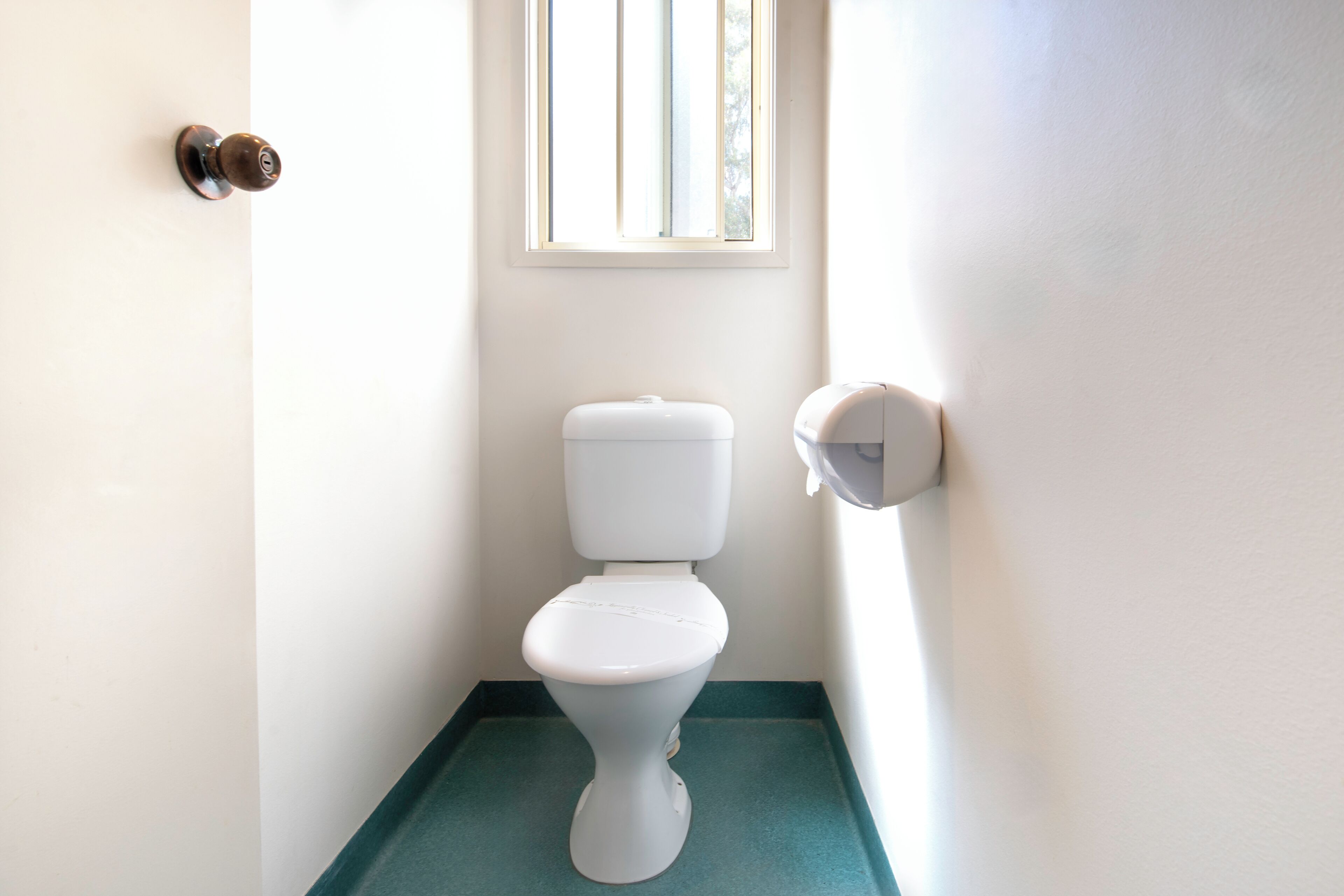 De petites toilettes blanches, avec un sol vert, une fenêtre au-dessus et un distributeur de serviettes en papier sur le côté droit.