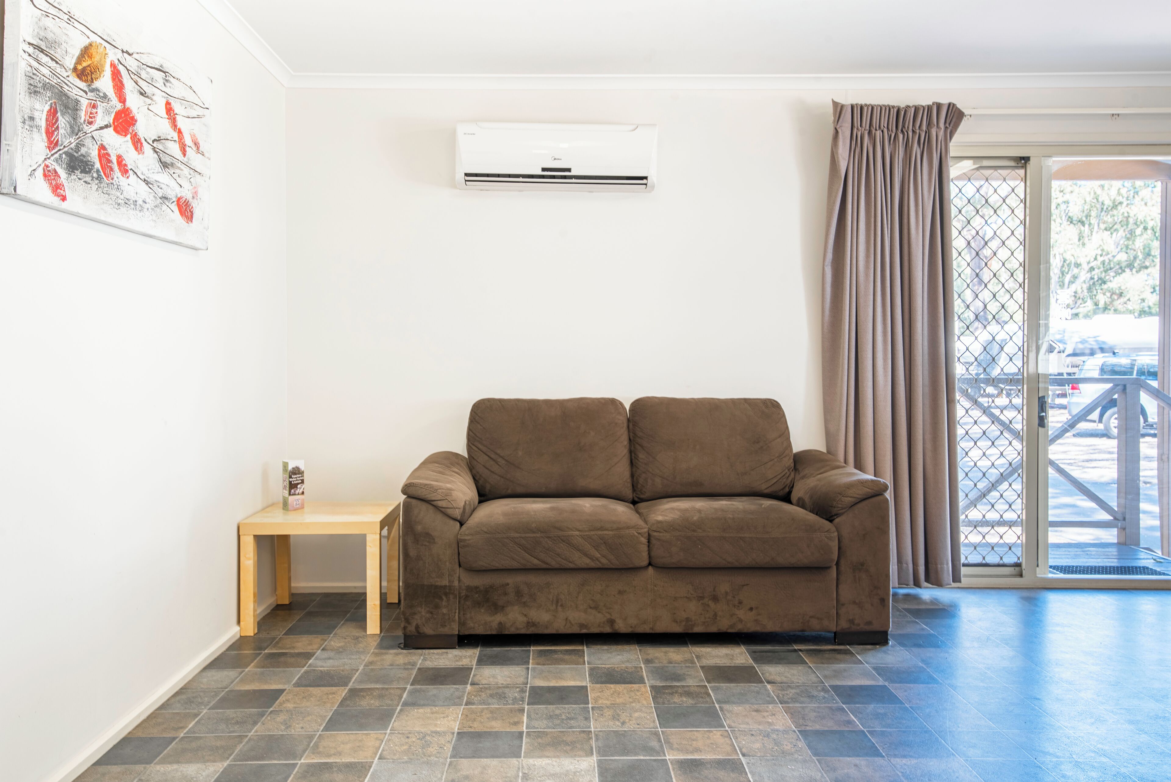 A living room with a brown sofa, a small wooden table, a white air conditioning unit, and a glass door leading outside.