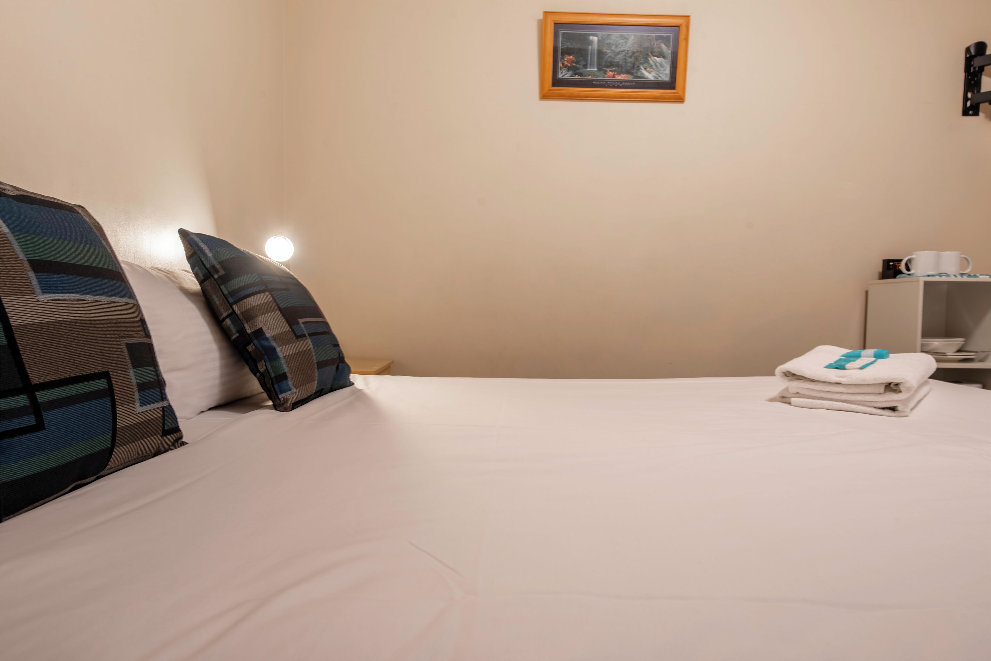 Una cama bien hecha con almohadillas a rayas, un cuadro en la pared y una pila de toallas dobladas en la mesita de noche.