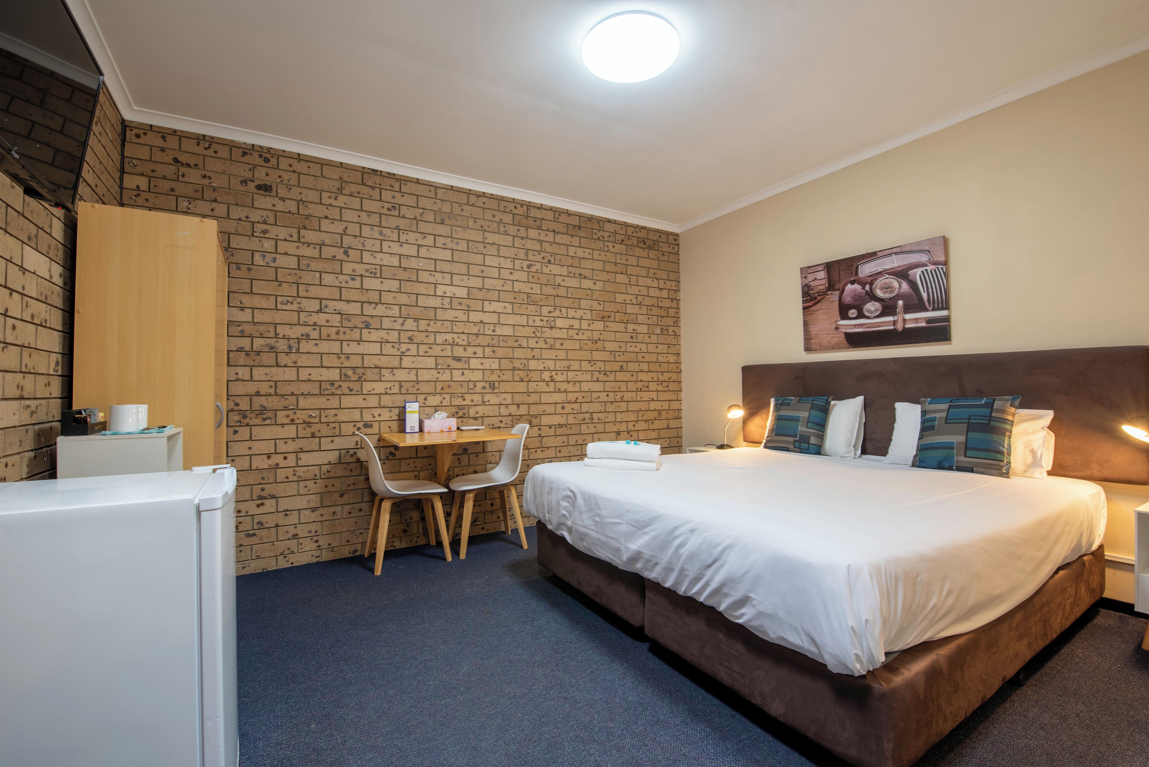 Habitación de hotel con pared de ladrillo, cama con ropa blanca, escritorio y dos sillas, y un cuadro en la pared.