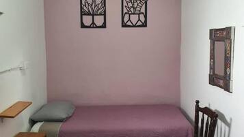 Una habitación pequeña con cama individual, pared rosa, mesita de noche de madera, una silla de madera y dos cuadros enmarcados.