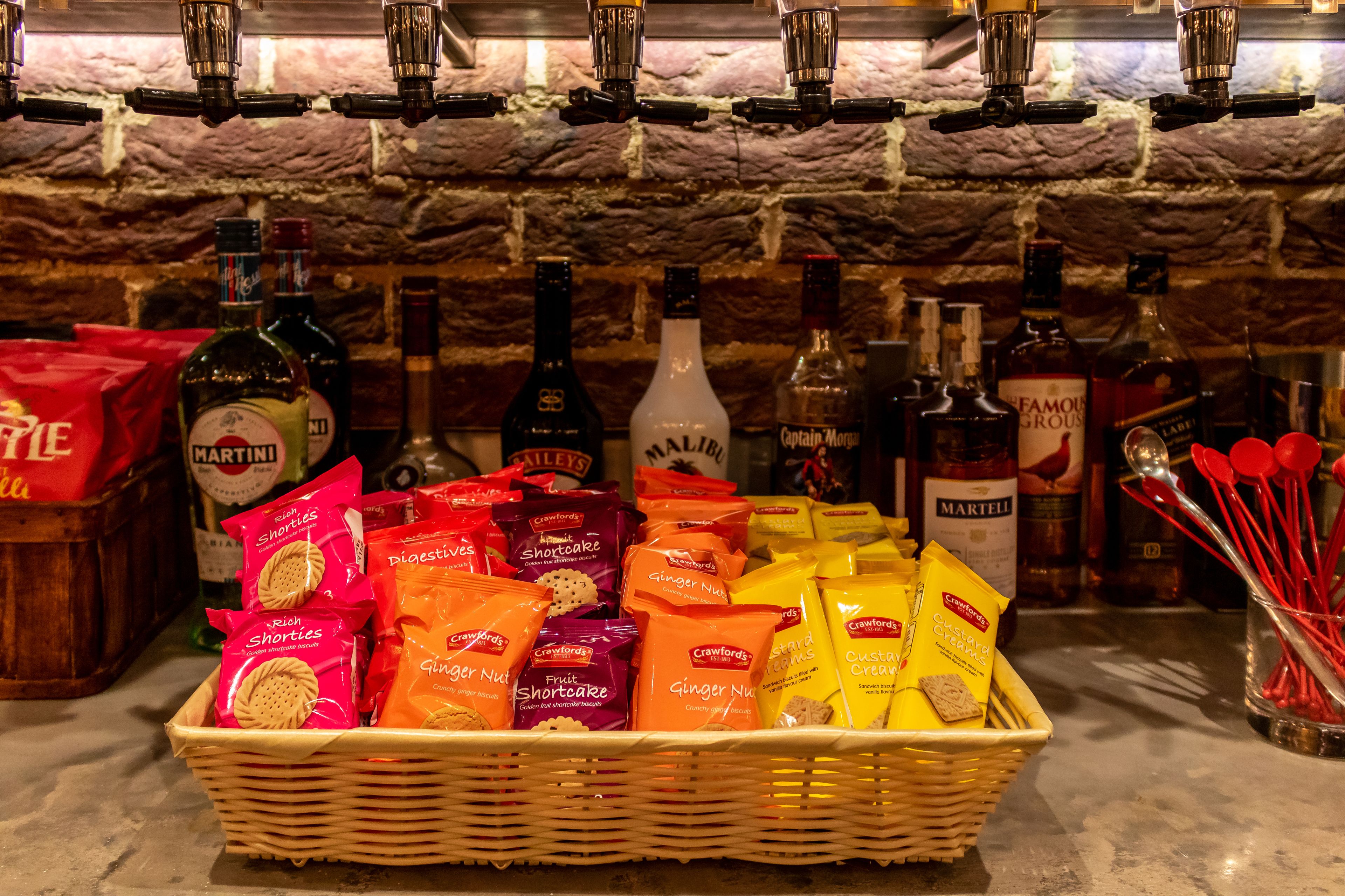 Een bar met diverse drankflessen, een mand vol snacks en een achtergrond van een stenen muur.